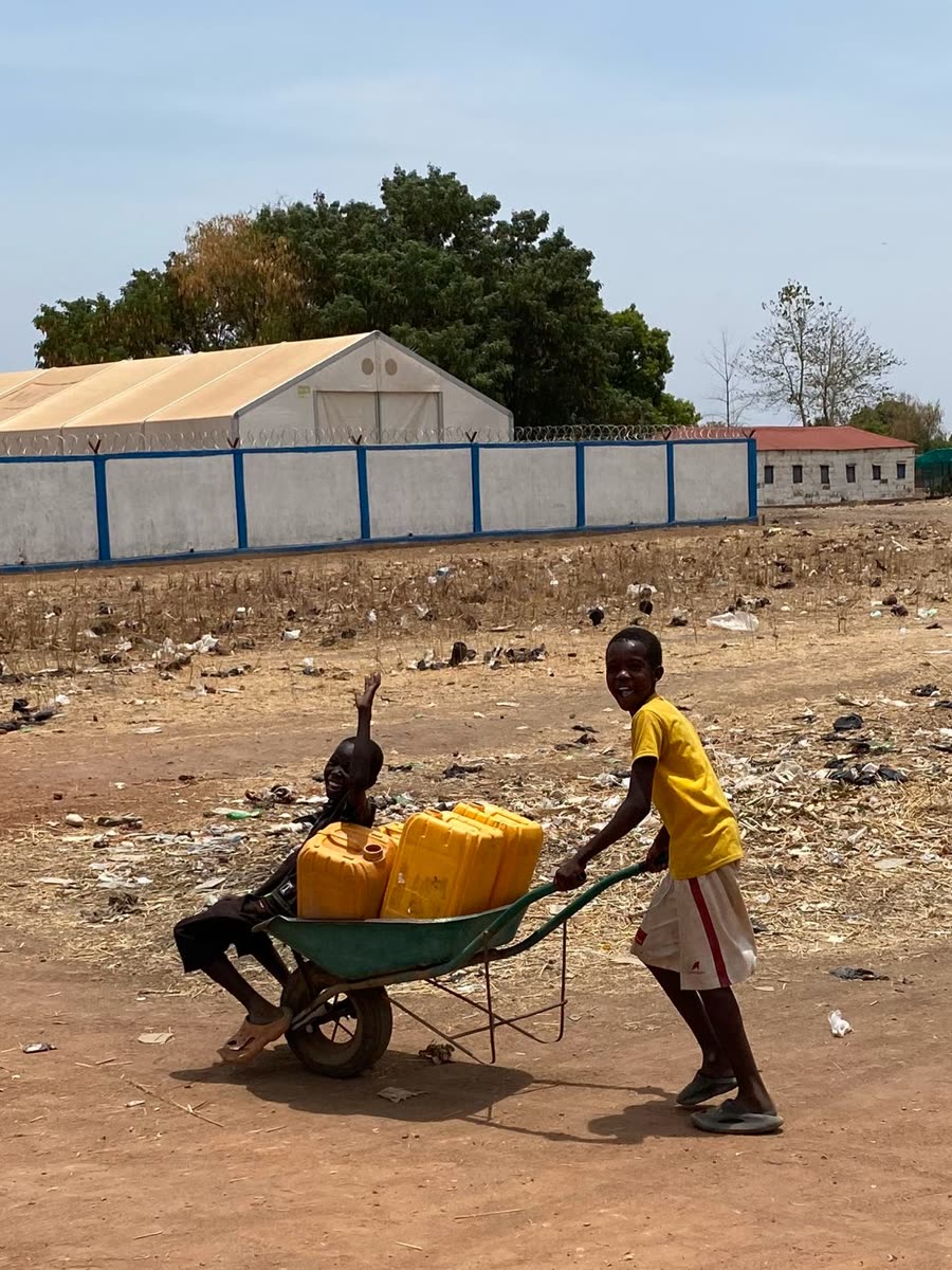 Deux enfants souriants poussant une brouette chargée de bidons d'eau près d'un camp